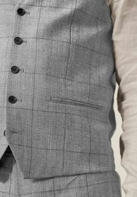 Gilet grigio a quadri con bottoni neri indossato sopra una camicia a maniche lunghe beige chiaro, che mostra pantaloni grigi a quadri coordinati.