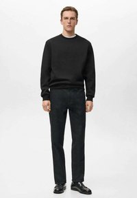 Čierna mikina s rebrovaným okrúhlym výstrihom, kombinovaná so vzorovanými tmavosivými nohavicami s rovnými nohavicami. Model má na sebe čierne kožené topánky a stojí na bielom pozadí.