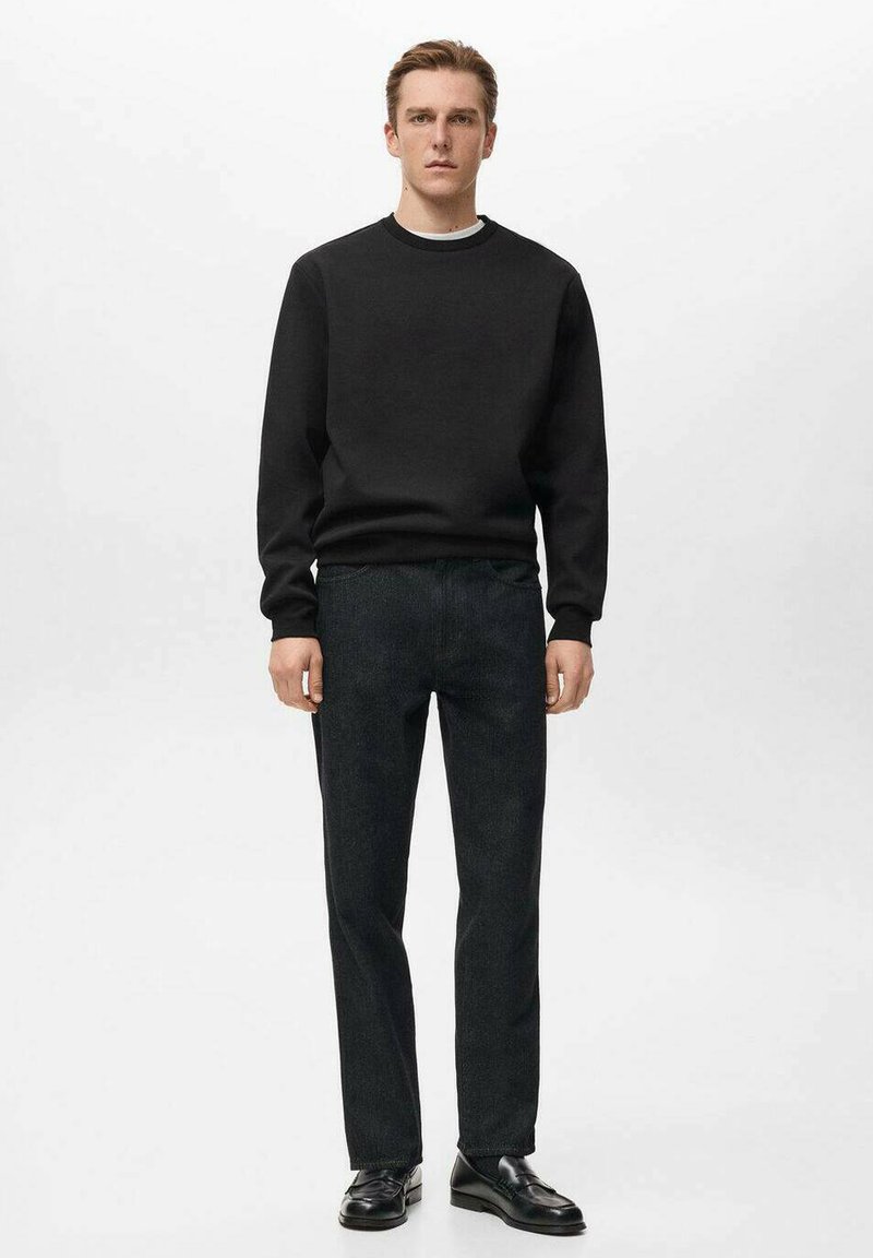 Sudadera negra con cuello redondo acanalado, combinada con pantalones rectos de un gris oscuro. El modelo lleva zapatos de cuero negros y está de pie frente a un fondo blanco.