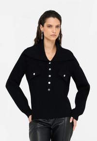 Maglione nero a coste con colletto ampio, dotato di due tasche frontali e bottoni argentati lungo la patta. Tessuto in maglia testurizzata.