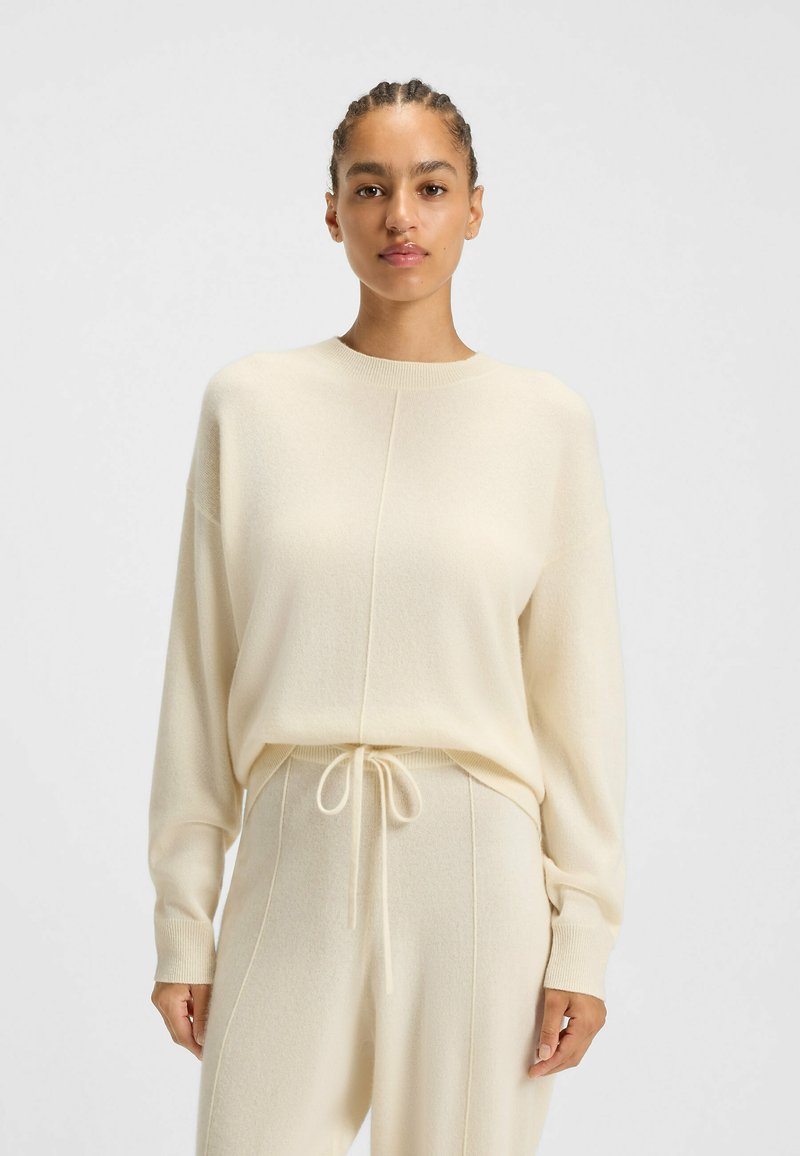 Helle beigefarbene Strickjacke aus weichem, strukturiertem Stoff mit rundem Ausschnitt. Verfügt über überschnittene Schultern und einen lässigen Schnitt. Passende Jogginghosen mit Kordelzug.
