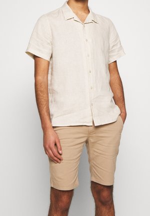 Mann trägt ein beigefarbenes Hemd mit kurzen Ärmeln und Kragen sowie beige knielange Shorts, steht mit einer Hand in der Tasche.