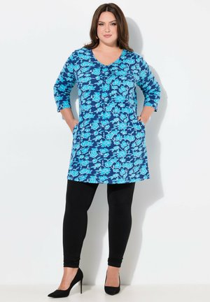 Blaue Blumentunika mit V-Ausschnitt und langen Ärmeln; verfügt über eine Knopfdetaillierung und zwei Seitentaschen, kombiniert mit schwarzen Leggings und Heels.