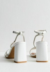 White heeled sandals feature a block heel, thin ankle straps adorned with silver accents, and a smooth leather texture.