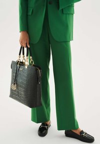 Musta teksturoitu käsilaukku, jossa on kullanväriset metalliosat, mustat nahkahihnat ja kultainen koriste, yhdistetty vihreään räätälöityyn pukuun ja mustiin loafereihin.