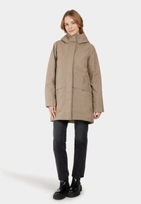 Beige wasserdichte Jacke mit Kapuze, langen Ärmeln, vorderem Reißverschluss und großen Fronttaschen, kombiniert mit schwarzen Jeans und robusten schwarzen Stiefeln.