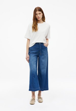 Jonge vrouw met lang bruin haar, een wit T-shirt met korte mouwen, wijde cropped blauwe spijkerbroek en beige schoenen, staande met één hand in haar zak.