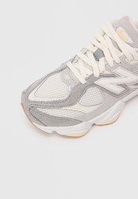 Baltos ir pilkos New Balance sportbačiai su tinklelio ir zomšo detalėmis, grietinėlės spalvos raišteliais ir smėlio spalvos akcentais ant pado, izoliuoti ant balto fono.