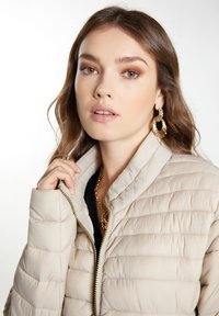 Giubbotto imbottito beige con texture a righe orizzontali, zip dorata e colletto rialzato. La modella tiene il colletto con un orecchino e una collana con texture.