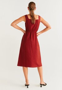 Femme aux cheveux bruns en queue de cheval portant une robe midi rouge sans manches et des sandales à talons noires, se tenant avec les mains sur les hanches, de dos.