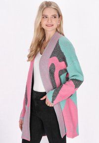 Cardigan colorato con motivi rosa, teal e grigio, dalla texture morbida, design aperto e bordi a costine, indossato su una maglietta bianca.