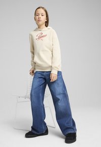 Felpa crema con logo Puma rosso, tasca frontale e polsini a coste. Jeans in denim blu a gamba larga e sneakers nere completano l'outfit.