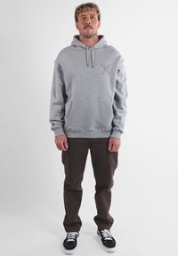 Felpa grigia con cappuccio e tasca frontale, caratterizzata da una piccola grafica colorata sul petto, abbinata a pantaloni marroni e sneaker nere.