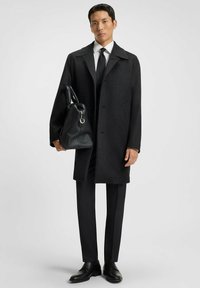 Cappotto a scacchi neri con una vestibilità sartoriale, abbinato a una cravatta nera e una camicia elegante. Tiene una borsa in pelle nera strutturata.
