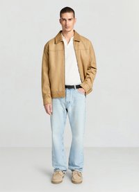 Veste en similicuir beige avec col, polo blanc à motifs, jean bleu clair et mocassins beiges. Homme debout, mains dans les poches.