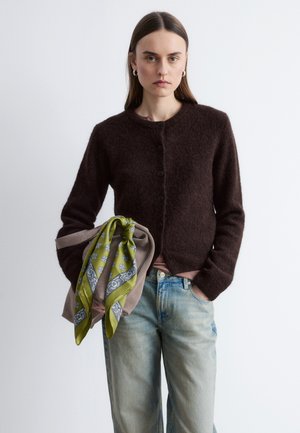 Vrouw draagt een donkerbruine, zachte cardigan, blauwe spijkerbroek, houdt een beige jas vast met een groene sjaal met patroon, staand tegen een effen achtergrond.