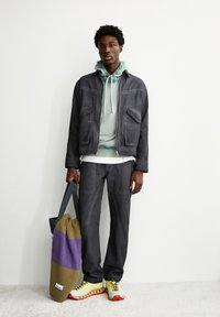 Dunkelgraue Denimjacke über einem mintgrünen Hoodie, kombiniert mit passenden grauen Hosen und gelben Turnschuhen. Eine mehrfarbige Tasche wird in einer Hand gehalten.