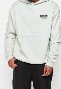 Sudadera ligera verde menta con un bolsillo frontal, puños elásticos y un pequeño parche negro con el logo que dice "ENFRENTAR LA REALIDAD, BEBÉ" en el pecho.