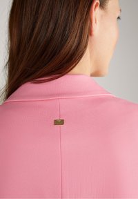 Nainen, jolla on pitkät ruskeat hiukset ja vaaleanpunainen takki, jonka selkäpuolella kauluksen alapuolella on pieni kullanvärinen logolevy.