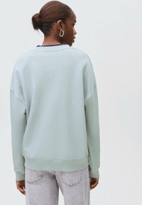 Zeleno pulover s sproščenim krojem, rebrastim robom in manšetami ter okroglo ovratnikom, nosi preko svetlo sivo oblečenih kavbojk z detajlom na zadnjem žepu.