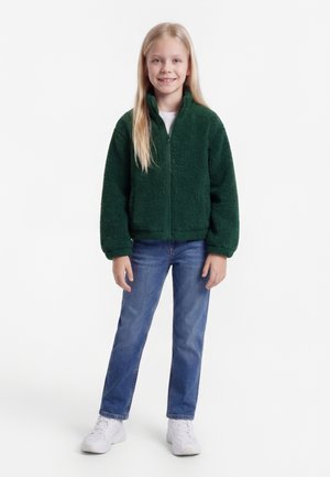 Blondínka stojí a usmieva sa, má na sebe tmavozelenú fleecovú bundu na zips, modré džínsy a biele tenisky na jednoduchom bielom pozadí.