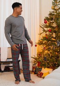 Grijs shirt met lange mouwen en rode geruite pyjamabroek met trekkoord. Staand naast een versierde kerstboom en cadeaus.