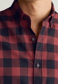 Chemise à carreaux rouge et marine avec col boutonné, boutons contrastants et détails de couture visibles. Le tissu semble doux et texturé.