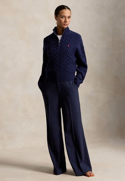 Nő tengerészkék patentos fonott mintás pulóverben és széles szárú tengerészkék nadrágban, zsebekben tartott kézzel, bézs háttér előtt áll.