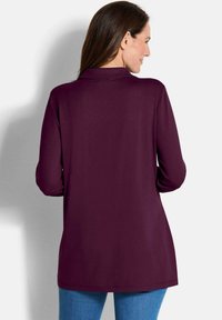 Tunika bordeaux con maniche lunghe, colletto e taglio dritto. Tessuto morbido, texture liscia e design all'altezza dei fianchi. Indossata con jeans blu.