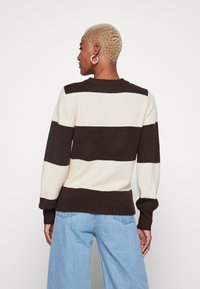 Pull rayé marron et crème avec des manches longues, ourlet et poignets côtelés, fabriqué en tissu tricot doux. Porté par-dessus un jean en denim bleu clair.