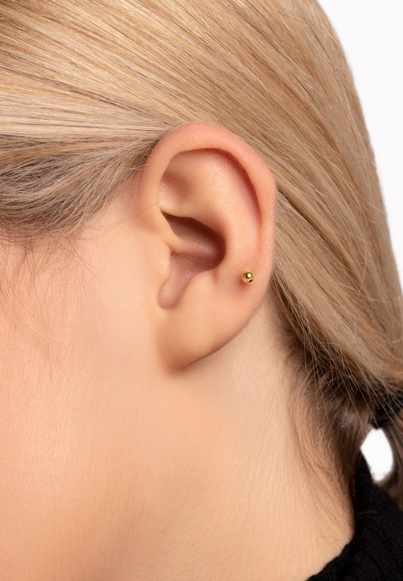 Orecchino a perno in oro posizionato sul lobo dell'orecchio, forma rotonda, superficie liscia, design minimalista, valorizzato da capelli biondo chiaro.