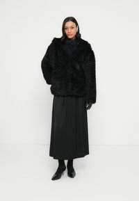 Czarna kurtka z futerkiem z imitacji, o luźnym kroju, z szerokim kołnierzem i długimi rękawami, zestawiona z czarną plisowaną spódnicą i butami na obcasie.