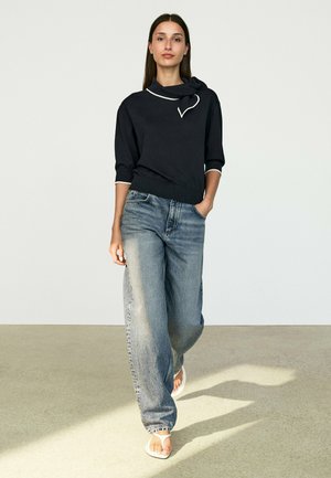 Donna che cammina all’interno indossando un top nero con rifiniture bianche, jeans blu larghi e scoloriti e sandali bianchi su un pavimento chiaro.