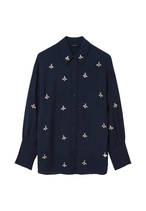 Camicia azzurra in denim con bottoni, realizzata in tessuto testurizzato, che presenta design di api ricamati in argento e bianco su tutta la parte anteriore e le maniche.