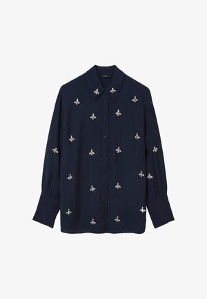 Camicia azzurra in denim con bottoni, realizzata in tessuto testurizzato, che presenta design di api ricamati in argento e bianco su tutta la parte anteriore e le maniche.