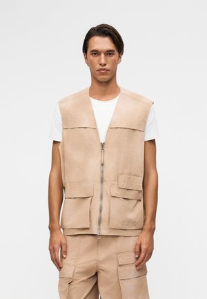 Man wearing beige sleeveless zip-up vest with multiple pockets over white short-sleeve shirt, matching beige cargo pants, standing against plain background.