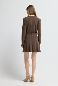 Abito marrone con corpino aderente, gonna svasata e vita strutturata. Indossato con scarpe beige traforate con tacco alto. Linee semplici e pulite.