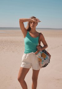 Tyrkysový top na ramínka, bílé pruhované šortky a barevná taška s vzorem geometrických tvarů a slunečními motivy, položeny na písčité pláži.