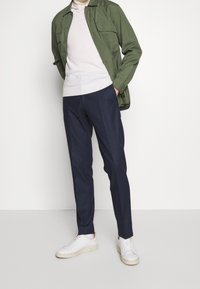 Giacca button-up verde oliva sopra un maglione leggero, abbinata a pantaloni a quadri blu navy e sneakers bianche. Taglio semplice e moderno.