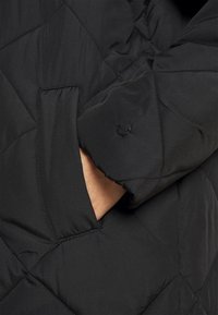 Svart vadderad pufferjacka med synlig ficka och knäppning. Materialet är slätt med en något glänsande yta.