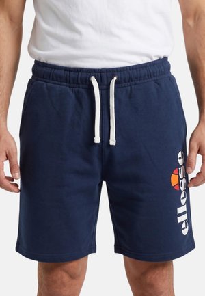Homme portant un short bleu marine avec cordon blanc et logo "ellesse", assorti d'un t-shirt blanc uni, debout devant un fond uni.