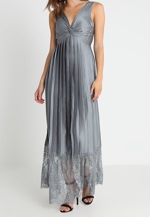 Vrouw draagt zilveren mouwloze geplooide maxi-jurk met kanten zoom en zwarte sandalen met hoge hak, staand tegen een witte achtergrond.