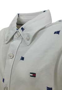 Polo shirt bianco realizzato in tessuto texturizzato, con piccoli motivi di barche a vela navy e un accento di marca vicino all'orlo con dettagli in rosso e blu.