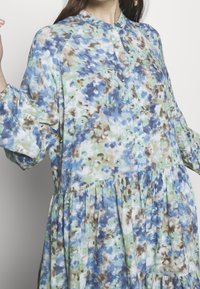 Blouse fleurie en tissu doux et léger présentant des tons bleus, verts et bruns. Col montant, ouverture à boutons sur le devant, et manches longues bouffantes avec un design à étages.