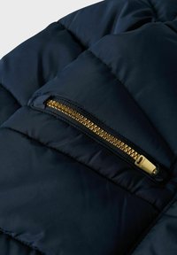 Navy puffer jacket section with a gold zipper pocket. Features quilted texture and smooth fabric, highlighting the contrast between colors.