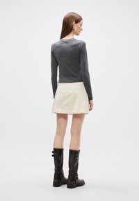 Γκρι μακρυμάνικο τοπ, κρέμα φούστα με πτυχές στο πλάι και μαύρες δερμάτινες μπότες μέχρι το γόνατο με πόρπες. Μοντέλο βλέπεται από πίσω.