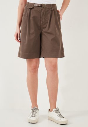 Personne portant un short marron ceinturé au genou et des baskets blanches, debout devant un fond clair uni.