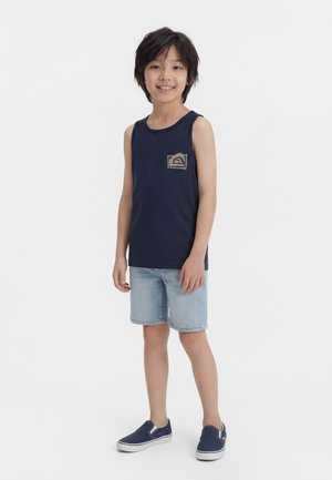 Dreng med mørkt hår iført en navy blå ærmeløs skjorte, lyse denimshorts og navy slip-on sko, der står og smiler mod en hvid baggrund.