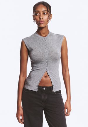 Femme portant un haut gris sans manches à boutons, partiellement déboutonné en bas, associé à un jean noir, debout devant un fond uni.