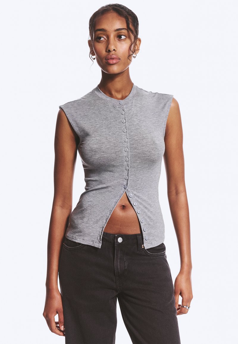 Femme portant un haut gris sans manches à boutons, partiellement déboutonné en bas, associé à un jean noir, debout devant un fond uni.